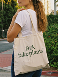 Person holding a tote bag with 'f*ck fake plants' text outdoors.